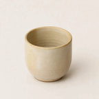 A small handmade ceramic espresso cup with a textured, speckled beige glaze, photographed against a solid, warm off-white background.