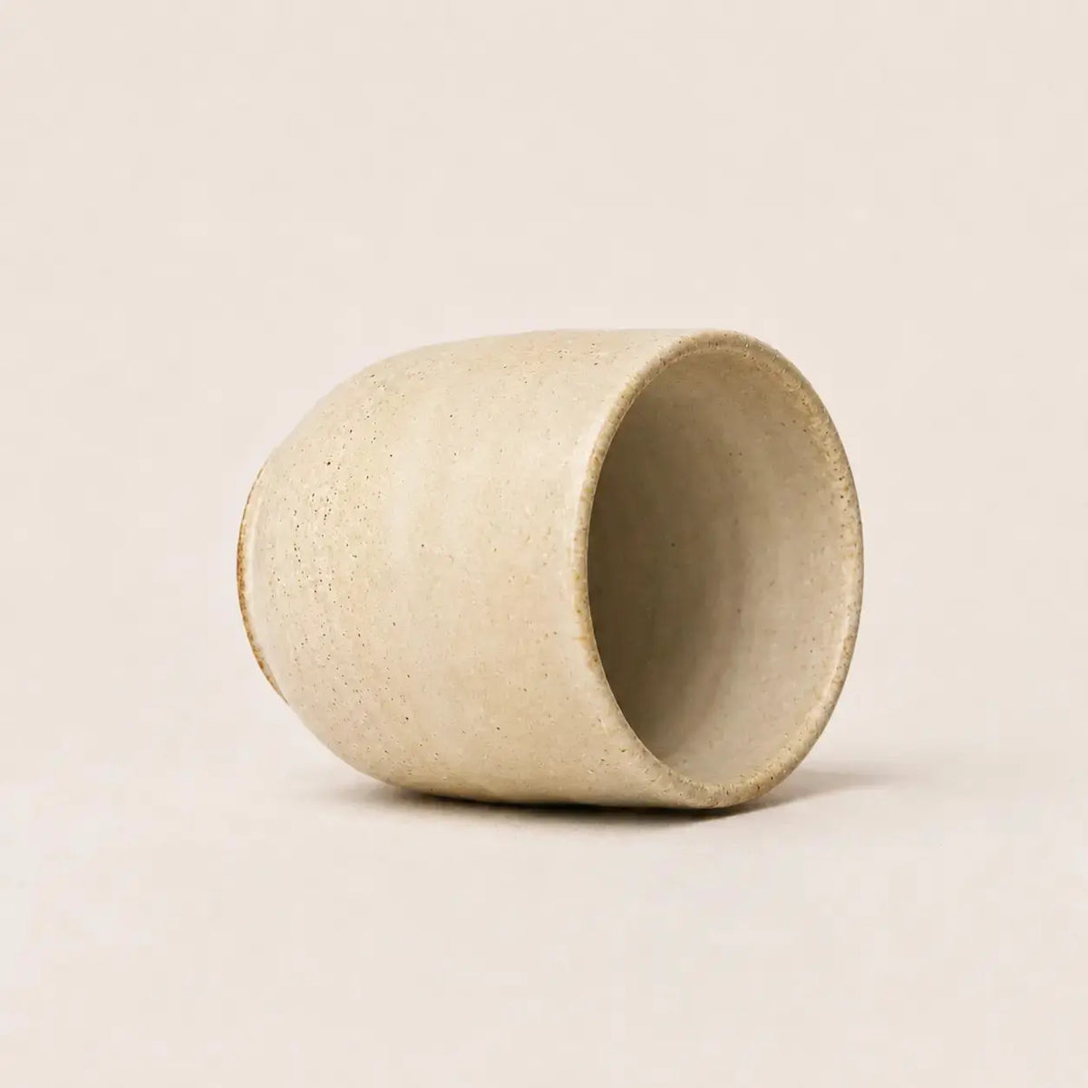 A small handmade ceramic espresso cup with a textured, speckled beige finish, shown on a plain, warm off-white background.