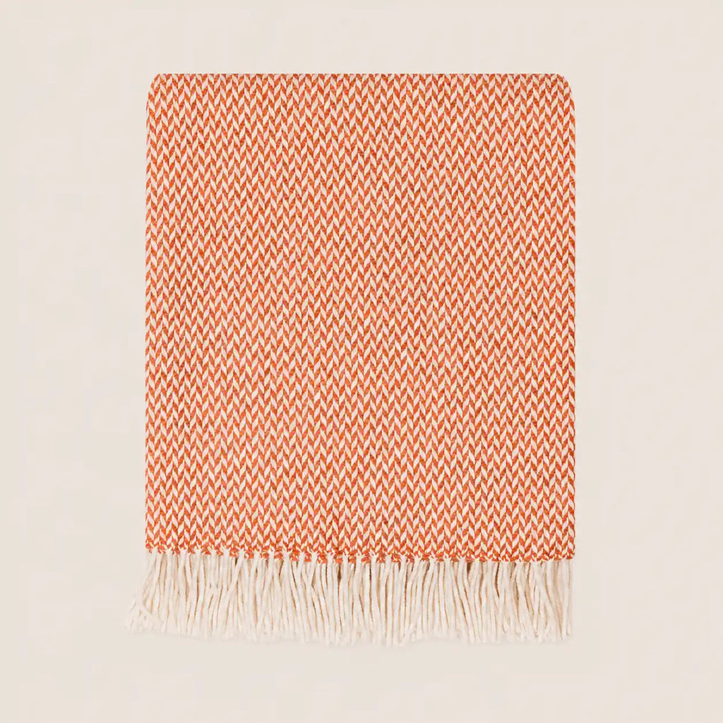 A folded woven throw blanket with a warm orange and white chevron pattern and fringed edges, displayed on a light beige background.