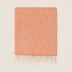 A folded woven throw blanket with a warm orange and white chevron pattern and fringed edges, displayed on a light beige background.