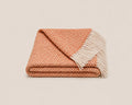 A folded woven throw blanket with a warm orange and cream chevron pattern and white fringe tassels, shown on a light beige background.