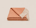 A folded woven throw blanket with a warm orange and cream chevron pattern and white fringe tassels, shown on a light beige background.