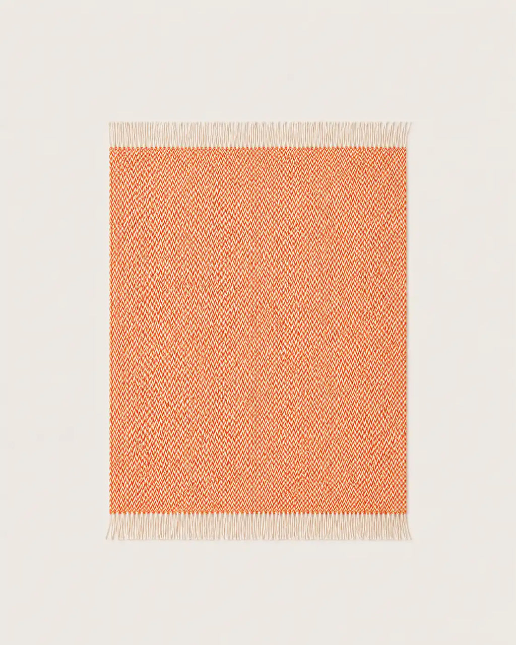 A woven throw blanket with a warm orange and cream chevron pattern and white fringe tassels, shown on a light beige background.