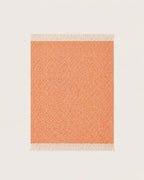 A woven throw blanket with a warm orange and cream chevron pattern and white fringe tassels, shown on a light beige background.
