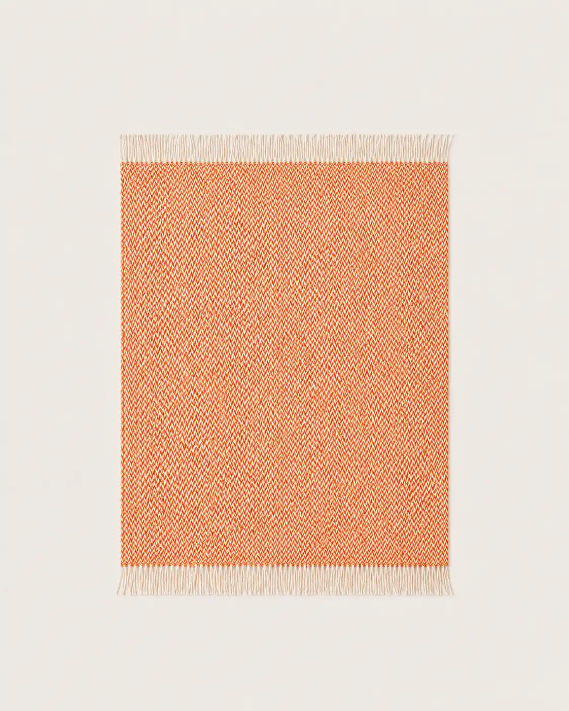 A woven throw blanket with a warm orange and cream chevron pattern and white fringe tassels, shown on a light beige background.