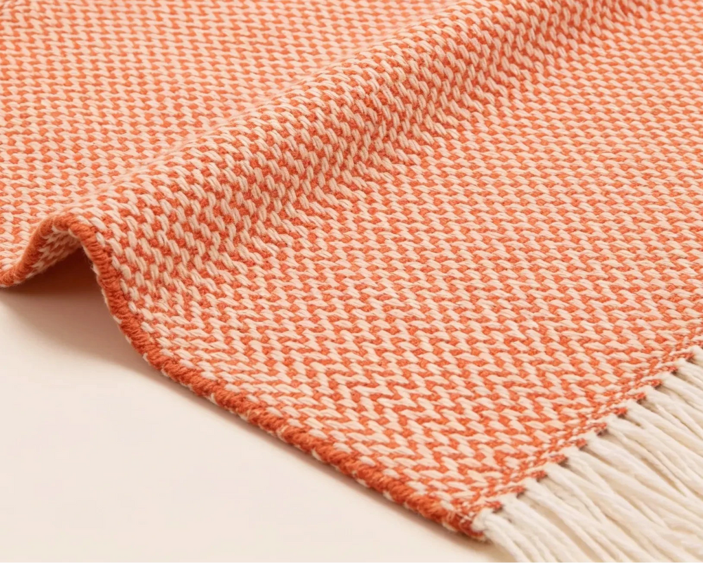A close-up of a woven throw blanket with a coral-orange and cream herringbone pattern and white fringe on one end, laid on a light neutral background.