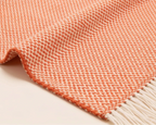 A close-up of a woven throw blanket with a coral-orange and cream herringbone pattern and white fringe on one end, laid on a light neutral background.