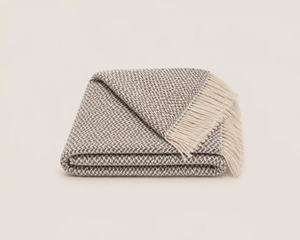A folded woven throw blanket with fringe, featuring a textured gray and white herringbone pattern, set against a solid off-white background.