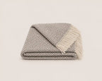 A folded woven throw blanket with fringe, featuring a textured gray and white herringbone pattern, set against a solid off-white background.