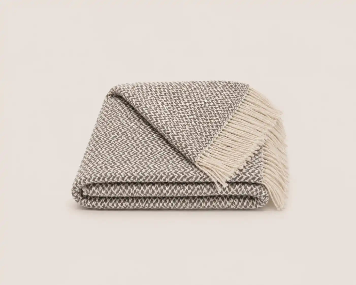 A folded woven throw blanket with fringe, featuring a textured gray and white herringbone pattern, set against a solid off-white background.