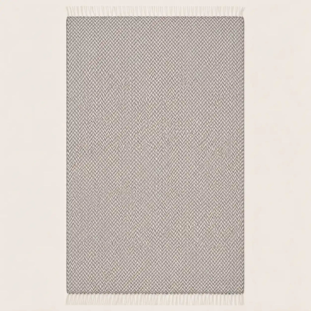 A rectangular throw blanket with a light gray and white textured herringbone pattern and white fringe on the top and bottom edges, shown against a plain off-white background.