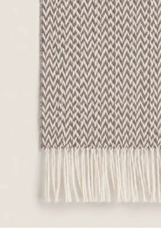 A close-up of a woven throw blanket with a small-scale herringbone pattern in taupe and off-white, finished with cream-colored fringe on both ends.