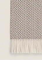 A close-up of a woven throw blanket with a small-scale herringbone pattern in taupe and off-white, finished with cream-colored fringe on both ends.