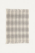 A rectangular woven throw blanket with a plaid pattern in beige, gray, and white, featuring fringed edges on the top and bottom, shown against a plain off-white background.