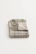 A folded woven throw blanket with fringed edges, shown on a plain white background. The blanket has a textured plaid pattern in off-white and gray.