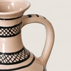Close-up of a ceramic pitcher with a glossy beige finish and black geometric lattice patterns, showing the handle and interior rim against a light neutral background.