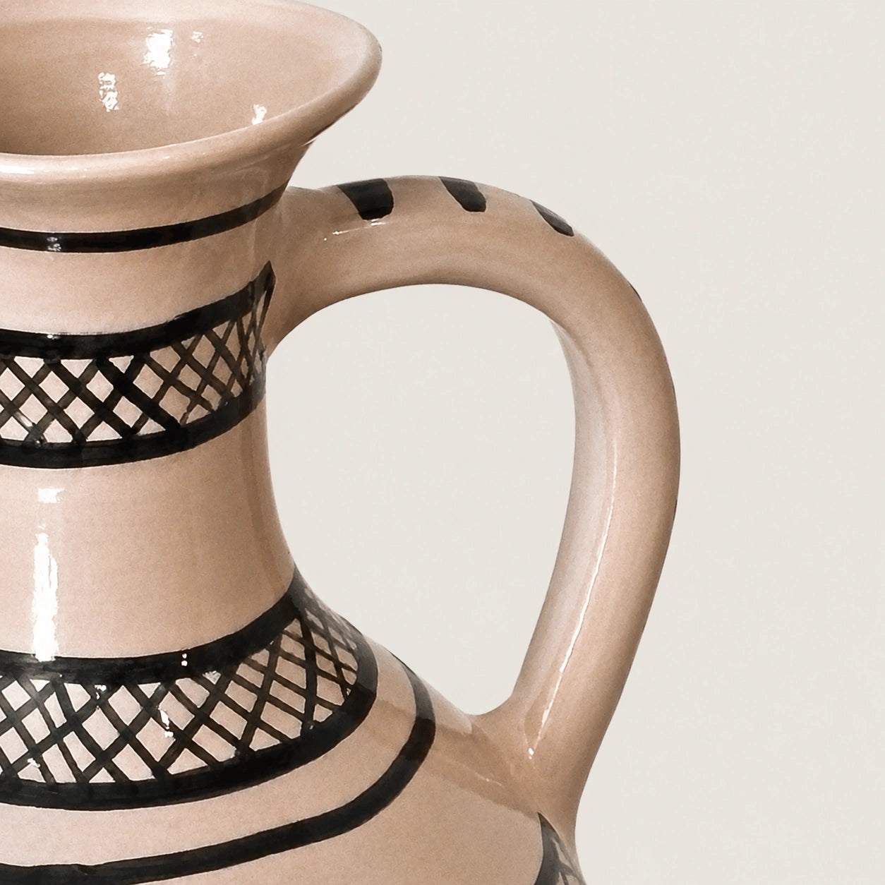 Close-up of a ceramic pitcher with a glossy beige finish and black geometric lattice patterns, showing the handle and interior rim against a light neutral background.