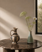 A decorative ceramic pitcher with black geometric patterns on a cream base sits on a dark marble table next to a light green vase holding dried flowers, set against a warm beige wall with soft window light.