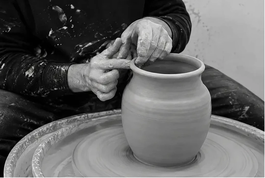 A black and white photo of a potter sitting at a pottery wheel, shaping a clay vase with their hands covered in clay dust.