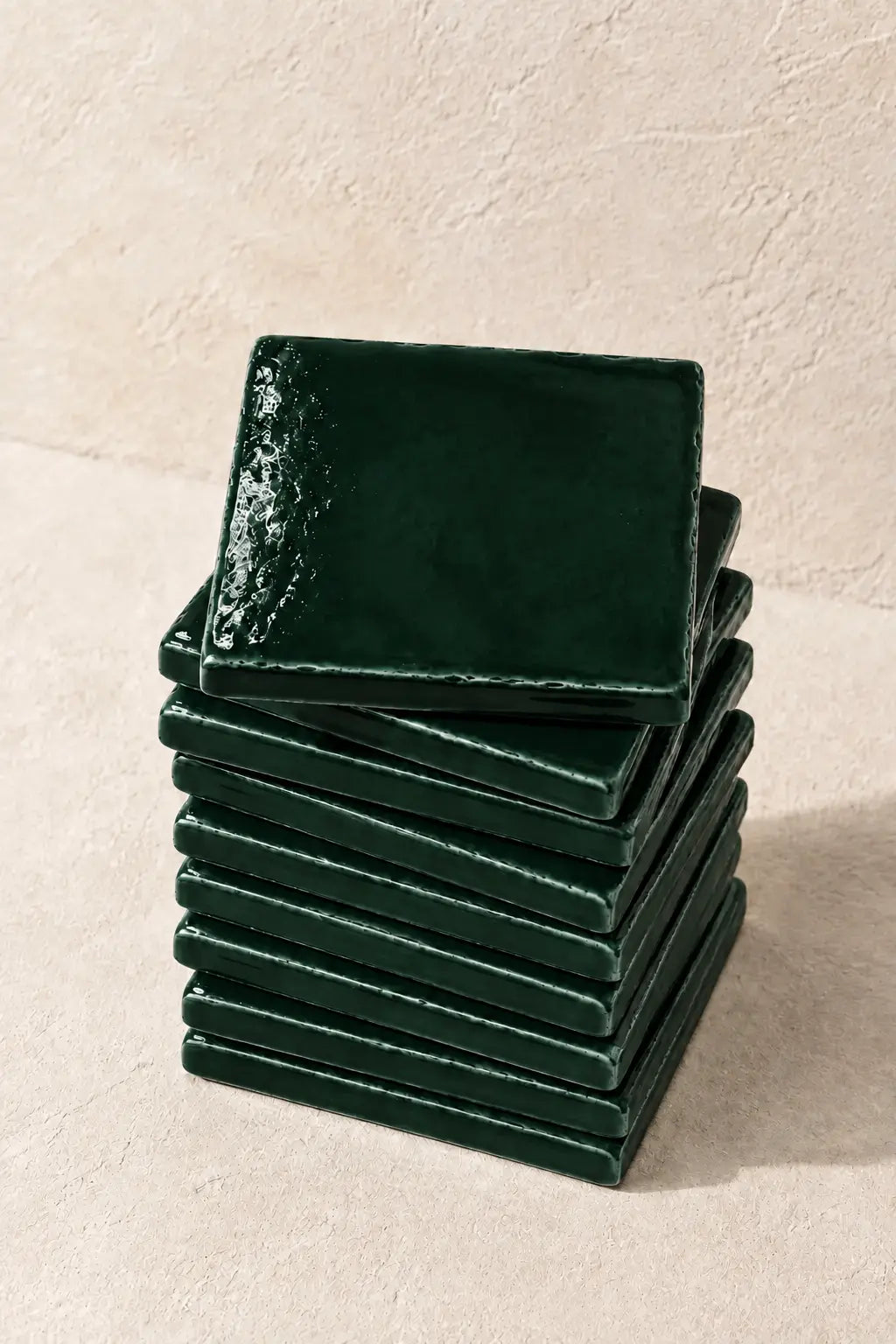 A stack of glossy forest dark green square coasters on a textured beige surface against a matching beige wall.