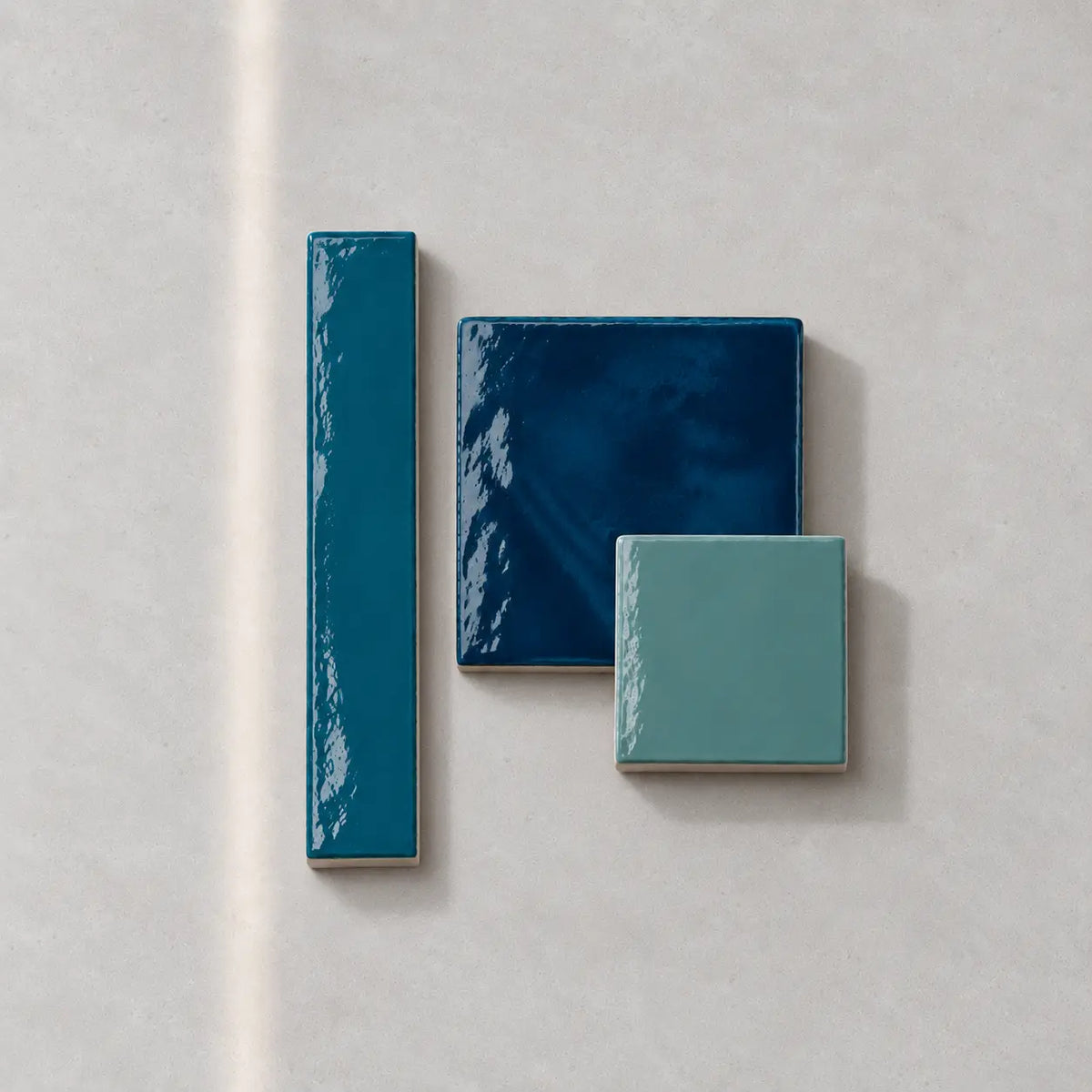 Three glossy ceramic tiles in shades of teal, dark blue, and light mint green, arranged on a light gray textured background.