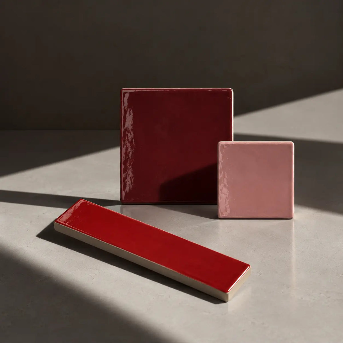 Three glossy ceramic tiles in red and dusty pink shades, arranged on a gray surface with dramatic diagonal shadows.