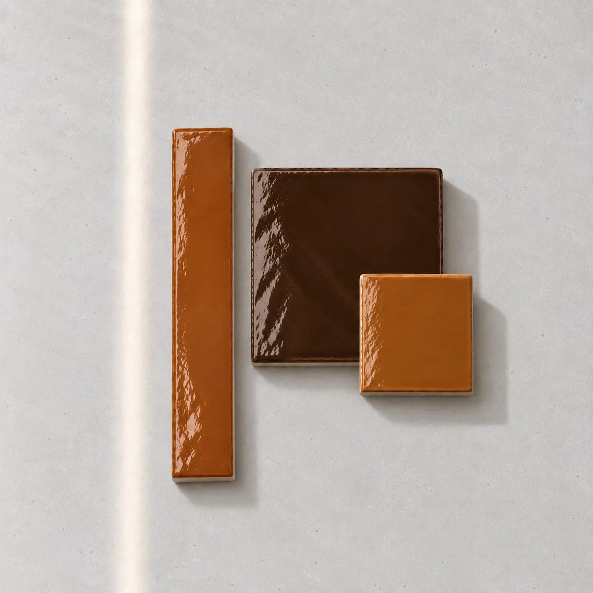 Three glossy ceramic tile swatches in brown and orange tones arranged on a light gray surface.
