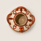 A top-down view of a round ceramic planter pot with a beige glaze and terracotta-colored handles and leaf-like patterns, set against a plain light background.