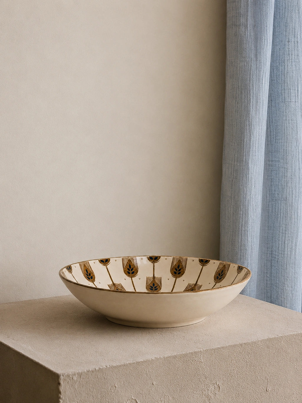 A shallow round ceramic bowl with a cream-colored glaze and a repeating brown tulip pattern around the rim, displayed on a textured stone pedestal against a neutral beige wall and light blue curtain.