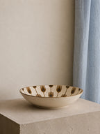 A shallow round ceramic bowl with a cream-colored glaze and a repeating brown tulip pattern around the rim, displayed on a textured stone pedestal against a neutral beige wall and light blue curtain.