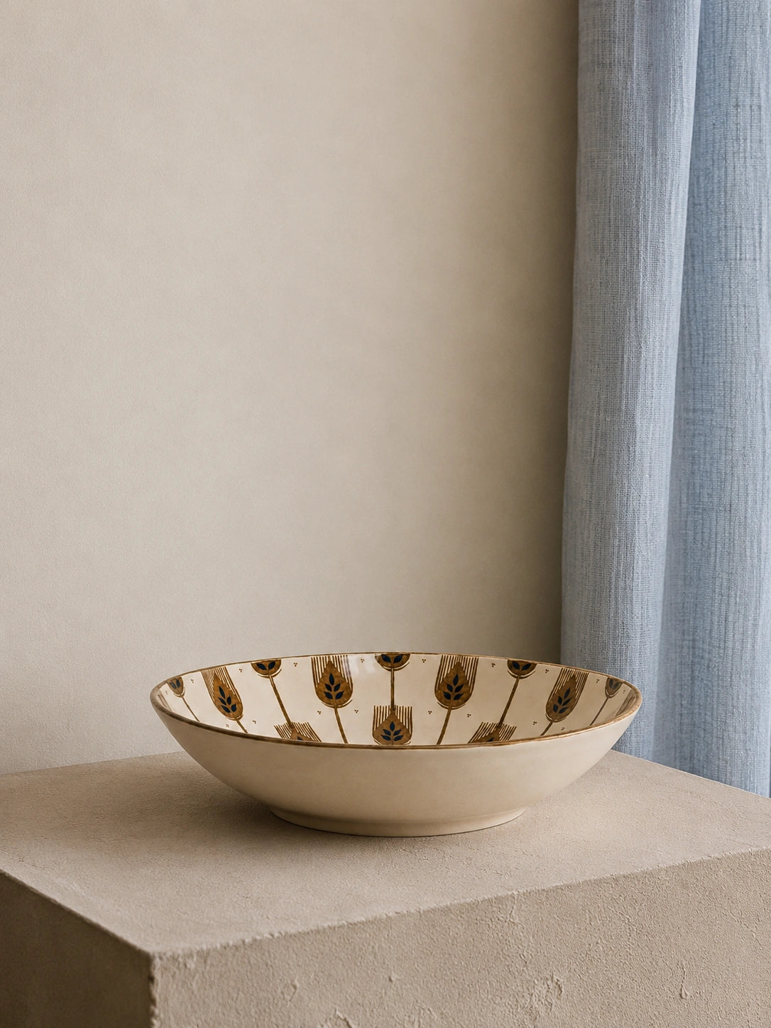 A shallow round ceramic bowl with a cream-colored glaze and a repeating brown tulip pattern around the rim, displayed on a textured stone pedestal against a neutral beige wall and light blue curtain.