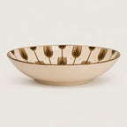 A shallow cream-colored ceramic bowl featuring a repeating pattern of stylized gold and dark blue-green botanical motifs on the interior rim, set against a plain light background.