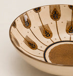 Close-up of the interior of a ceramic bowl with a warm cream glaze, decorated with metallic gold geometric and leaf-like motifs and small blue accents.