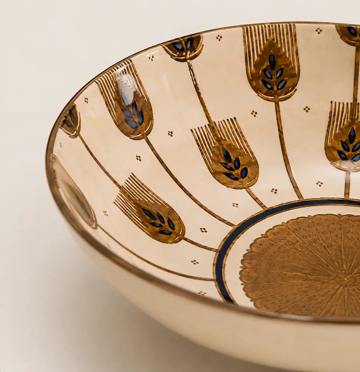 Close-up of the interior of a ceramic bowl with a warm cream glaze, decorated with metallic gold geometric and leaf-like motifs and small blue accents.