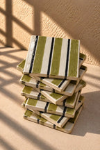 A stack of square ceramic tiles with a hand-painted green, white, and black stripe pattern, set against a textured beige stucco wall with warm sunlight and shadows.