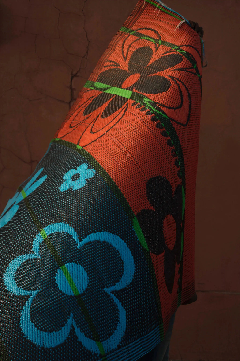 A close-up of a woven textile or rug featuring a bold floral pattern in orange, black, blue, and green, set against a dark brown textured background.