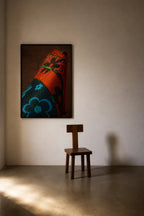 A framed abstract art print featuring a stylized red, black, and blue pattern hangs on a beige wall above a small wooden chair in a minimalist room with dramatic sunlight and shadows.