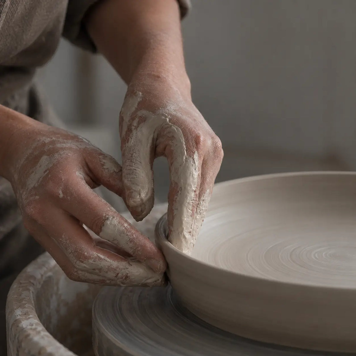 Close-up of a potter's hands covered in clay shaping a round ceramic plate on a pottery wheel.
