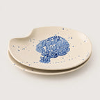 Set of two ceramic plates with a hand-painted blue tree design and blue speckles on a warm cream background, set against a light gray studio backdrop.