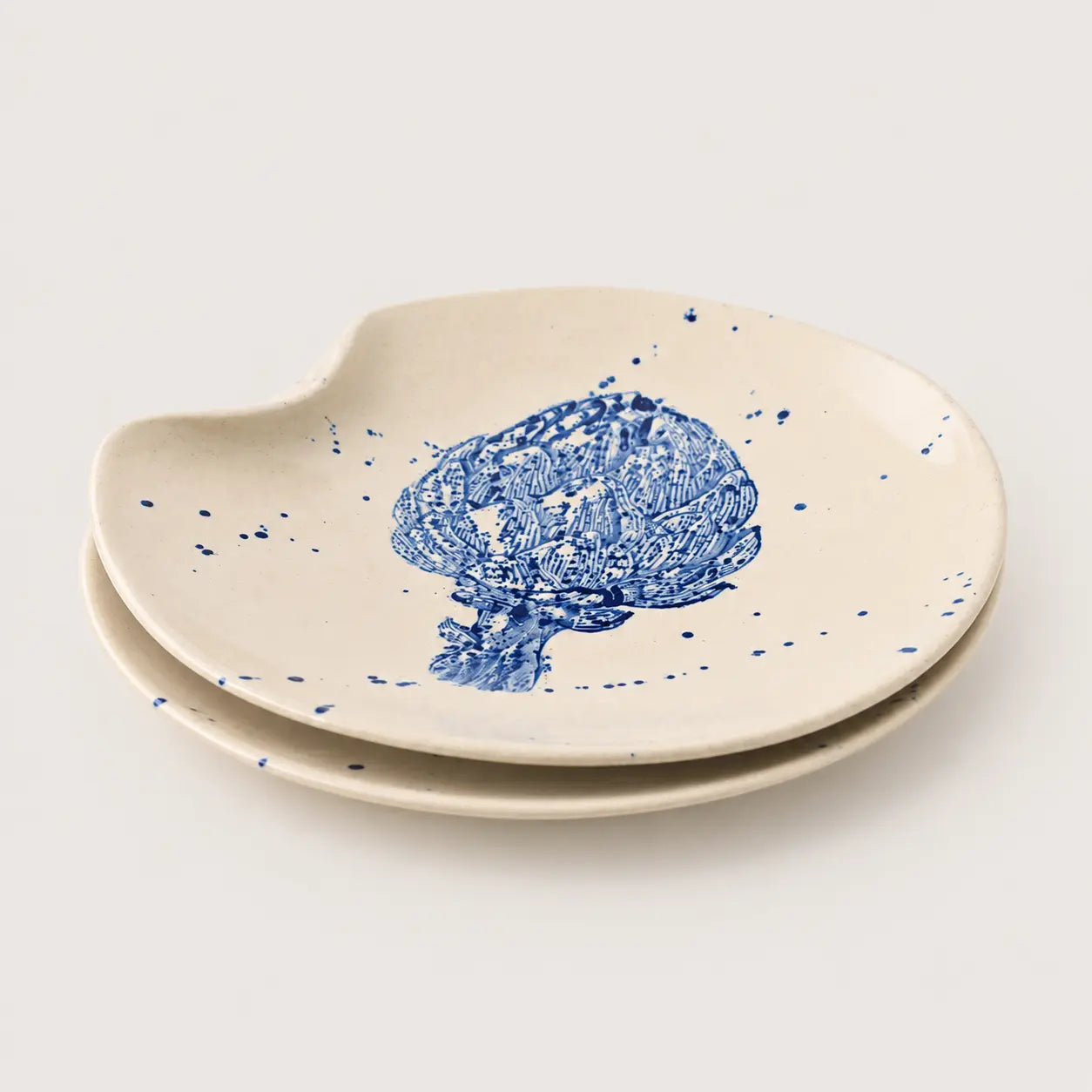 Set of two ceramic plates with a hand-painted blue tree design and blue speckles on a warm cream background, set against a light gray studio backdrop.