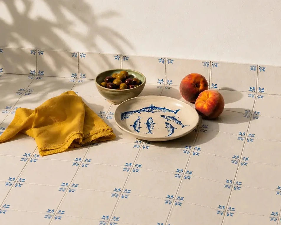 A styled tabletop scene with a white surface covered in a repeating blue geometric print, featuring a yellow cloth, a small bowl of olives, a plate with blue floral decoration, and two peaches under dappled sunlight.