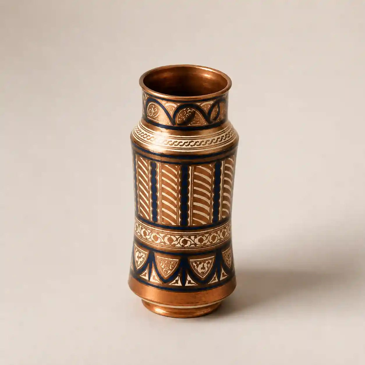 A small decorative vase with a warm metallic copper or bronze finish, decorated with dark navy blue and gold geometric patterns, set against a plain light beige background.