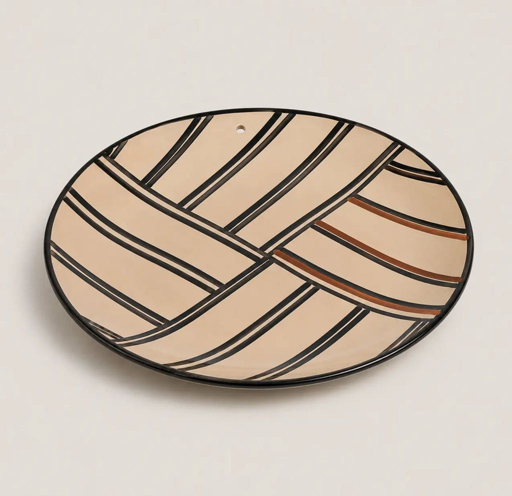 An oval ceramic plate with a geometric pattern of beige and cream stripes outlined in black, accented with thin brown lines, on a light beige background.