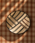 A round ceramic plate with a beige and brown basketweave pattern and a black rim, placed on a brown and beige checkered tablecloth with dappled sunlight.
