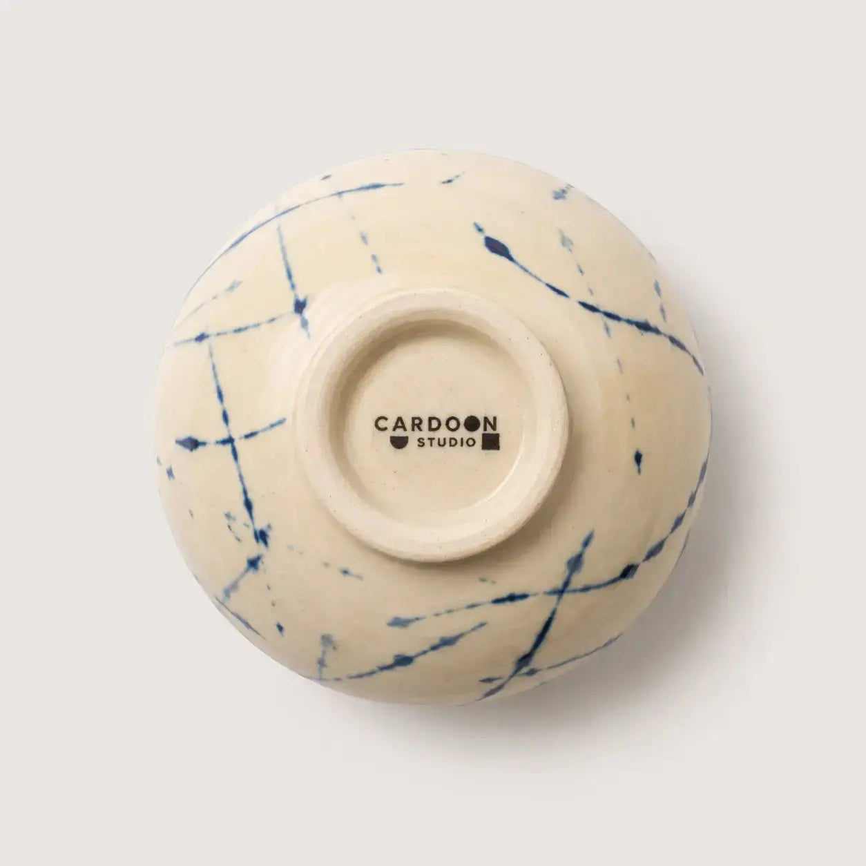 An off-white ceramic snack bowl viewed from underneath, featuring a blue ink-blot style pattern on the exterior and the brand name 'CARDON STUDIO' printed in black on the center base.