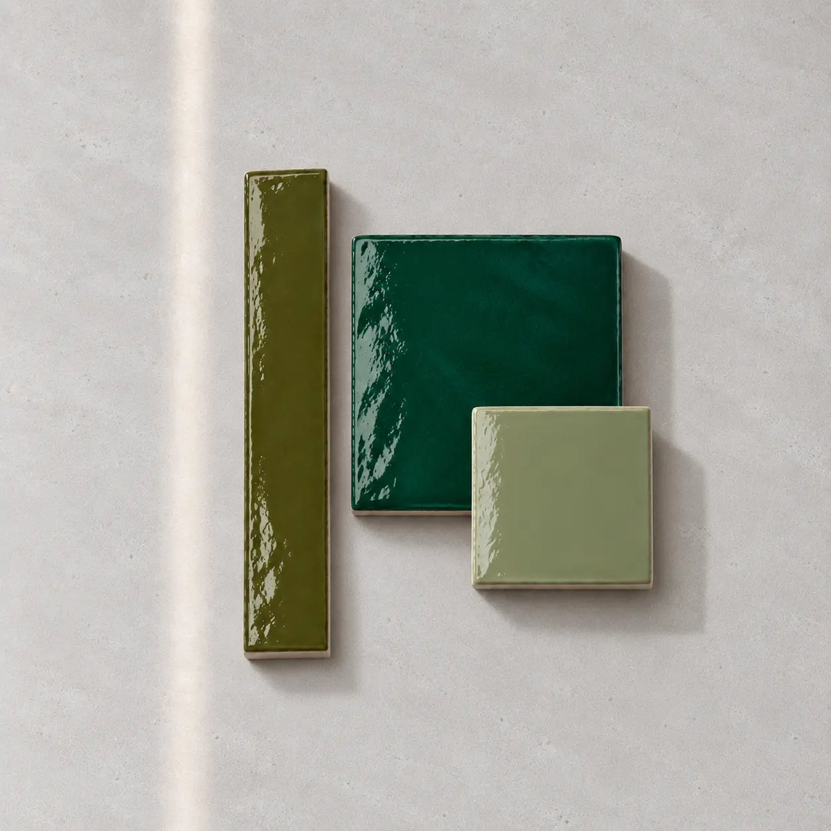 Three glossy ceramic tiles in olive green, dark forest green, and light sage green, arranged on a textured light gray surface.