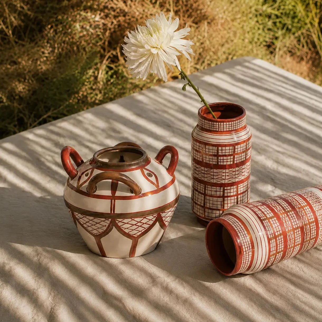 Three hand-painted ceramic vases in a terracotta and cream color scheme, styled on a beige linen cloth outdoors with dappled sunlight and a white flower in one vase.