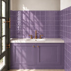A bathroom vanity setup with lilac paneled cabinets, a white countertop, an undermount sink, and a copper-toned faucet. The backsplash features glossy lilac square tiles, and a matching moss green tile wall surrounds the vanity.