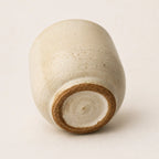 A handmade ceramic espresso cup with a rounded, slightly tapered shape, featuring a speckled off-white glaze and a contrasting brown clay rim at the base, photographed on a neutral light beige background.