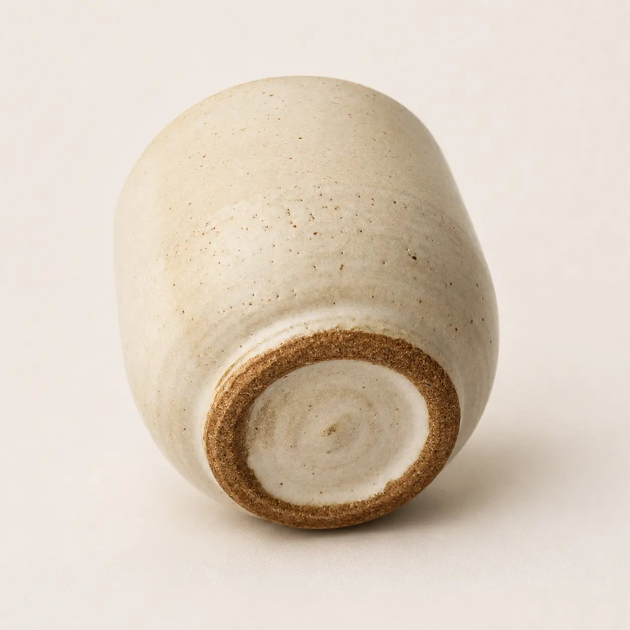 A handmade ceramic espresso cup with a rounded, slightly tapered shape, featuring a speckled off-white glaze and a contrasting brown clay rim at the base, photographed on a neutral light beige background.